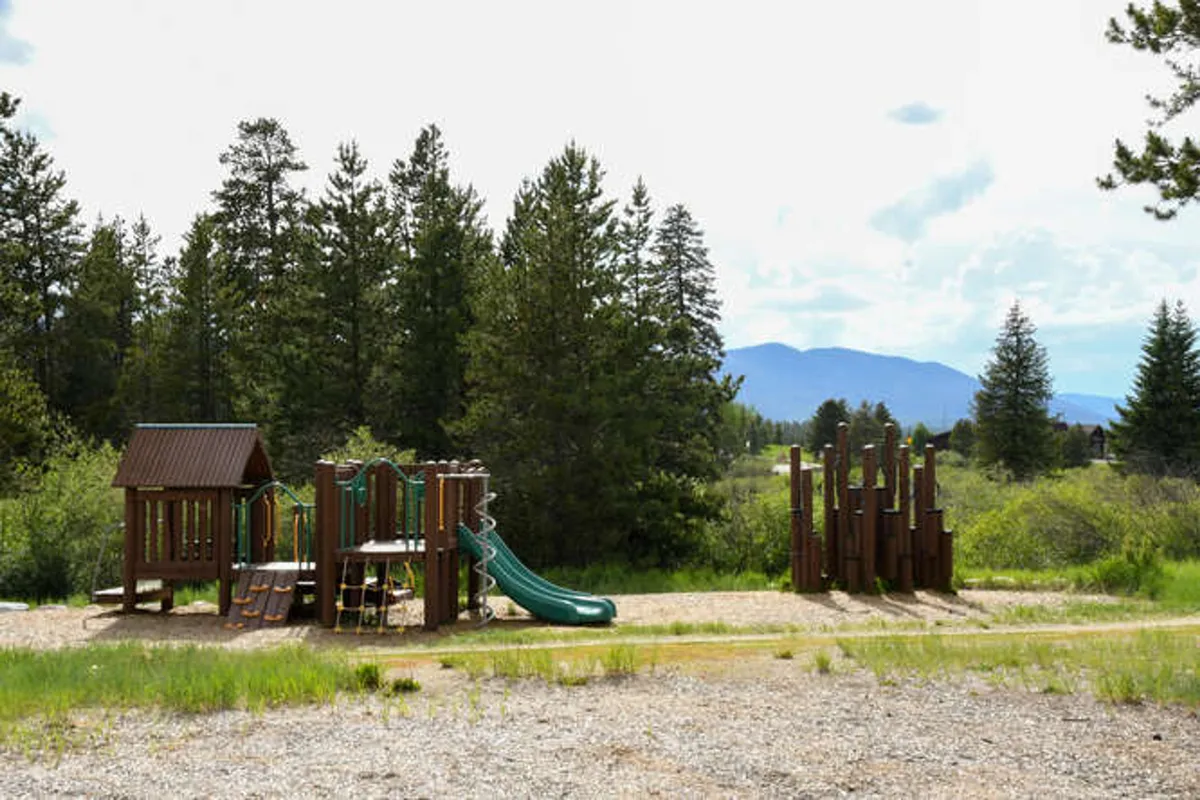 A joyful playground with slides, a tall wooden climbing structure, and nature all around, filled with children playing and families gathering. The vibrant, energetic atmosphere speaks to the nearby community amenities accessible from the home.