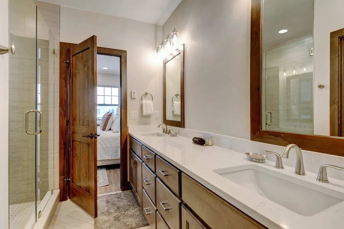 A spa-like bathroom showcasing a double sink vanity, large mirrors, a glass-enclosed shower, and wooden accents. The ambient lighting and spacious layout suggest comfort and relaxation, echoing the indulgence of spa-quality amenities.