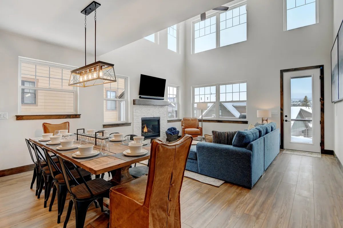 A bright, open living area showcasing a dining table perfect for group meals, a comfortable sofa, a modern fireplace, and oversized windows that look out onto the mountains. The ambiance is both cozy and elegant, designed for family gatherings or entertaining guests.