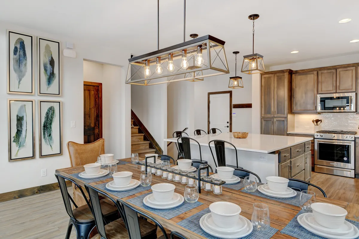 A warmly lit dining space with a custom wood dining table set for a meal, a striking chandelier above, and the adjacent kitchen island visible in the background. The room invites large group gatherings for dinners or games, and the open-concept layout keeps the kitchen and dining area connected.