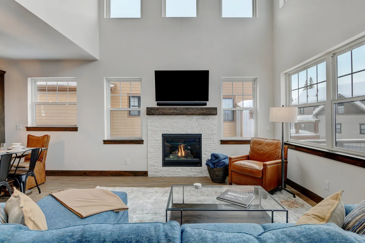 A spacious, welcoming great room featuring a cozy fireplace, comfortable seating, a large TV, and expansive windows that flood the space with natural light. The setting is ideal for gatherings, whether for movie nights, relaxed conversations, or group entertainment.