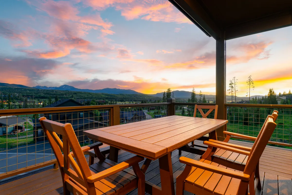 An outdoor scene featuring a wooden dining table and chairs positioned to take full advantage of sweeping mountain views and a colorful sunset sky. The setup suggests al fresco dining, relaxed conversations, and stargazing in a serene, resort-style environment.