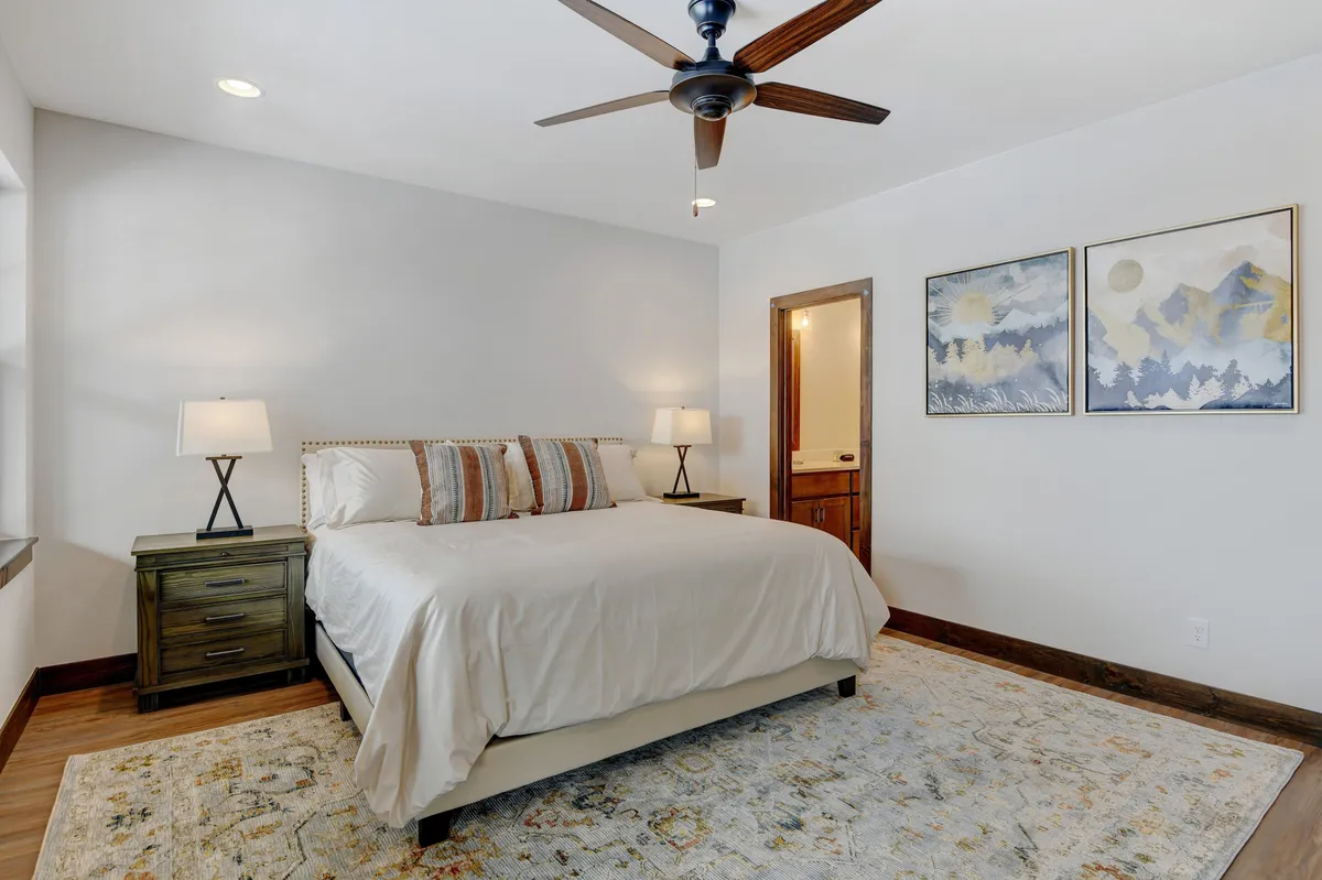 A tranquil bedroom featuring a plush bed with crisp white linens, matching nightstands with elegant lamps, decorative artwork above the headboard, and a soft rug at the foot of the bed. The space exudes calm and offers a restful atmosphere ideal for unwinding after an active day.