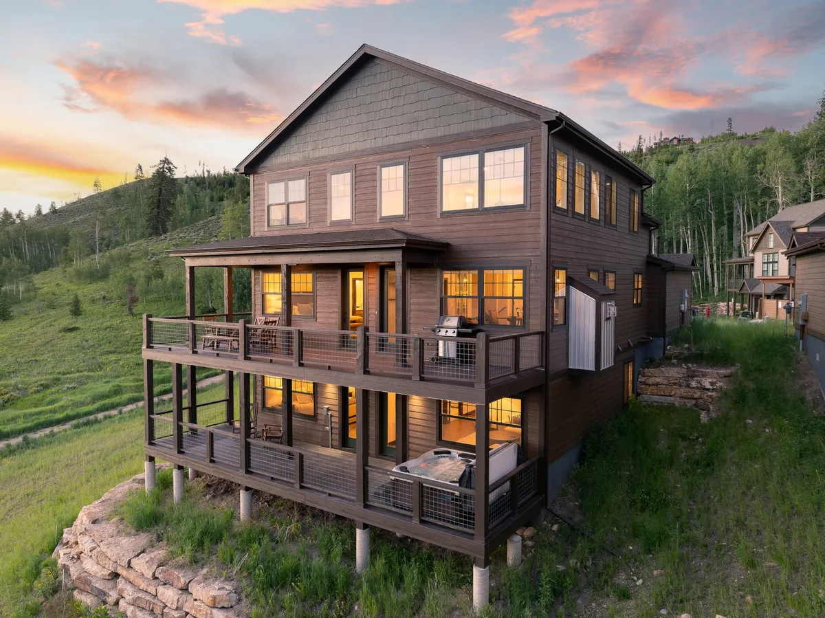 The image showcases the chalet's expansive wooden deck surrounded by lush greenery, with floor-to-ceiling windows reflecting the vibrant Colorado sunset. Guests can see an inviting outdoor dining area, a cozy fire pit perfect for gathering, and sweeping mountain views, setting the scene for relaxed evenings and nature-filled days.