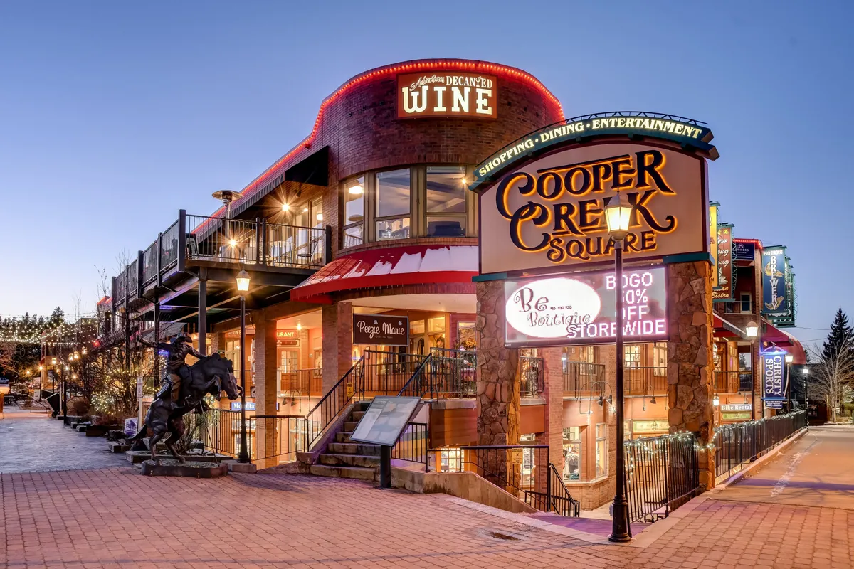 This exterior image features the recognizable Cooper Creek Square sign, a wine shop sign, stairs leading to an upper level, and a striking horse sculpture. The lively scene suggests a hub of activity perfect for groups to dine, shop, and enjoy entertainment together.